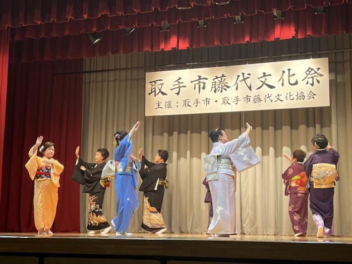 華やかな着物や落ち着いた色合いの着物を身にまとった複数の出演者が、舞台上で日本舞踊を披露しています