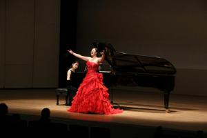 ホールの舞台で赤のドレスを着た女性が両手をあげ熱唱する様子。熱唱している女性の後ろに黒のドレスを着てピアノを弾く女性がいる。