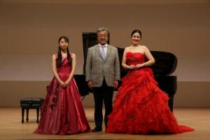 ドレスを着た2人の女性とスーツの男性がピアノの前に立っている