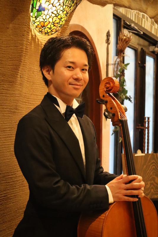 黒いスーツに黒の蝶ネクタイをした男性がチェロ片手に微笑んでいる写真