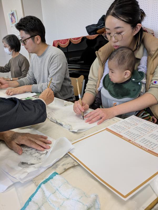 水墨画の体験教室にて、講師の丁寧な指導を受けながら、作品制作に取り組んでいる赤ちゃんを抱えた若いお母さんの様子