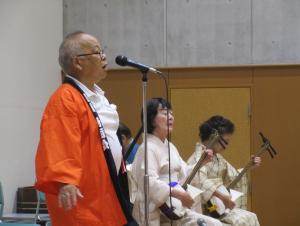 橙の法被を着た男性がマイクに向かい、画面奥では着物姿の女性2人が三味線を弾いている