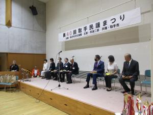 正装の男女８人が壇上の椅子に腰かけている。背面上方には「第４０回取手民謡まつり」の看板がかかっている
