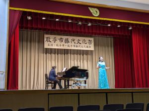 ステージで、ドレスの女性がピアノの伴奏で独唱している様子