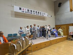 着物姿の男女数人が大勢の観客を前に舞台に立っている様子