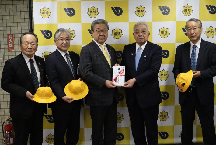 「ほどよく絶妙とりで」バックパネルの前で、黄色い帽子、目録を手に写真に写る5人の男性