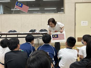 マレーシア国旗の前に立って話をする女性。その話を聞く子どもたちは、座ってメモをとっている。