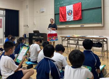 ペルー国旗の前に立って話をする女性。その話を聞く子どもたちは、座ってメモをとっている。