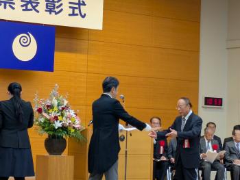 演題の前で正装の男性からスーツ姿の女性に賞状を手渡している