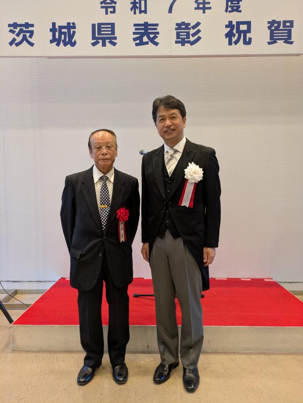 祝賀会上ひな壇の前での正装男性とスーツ姿男性のツーショット