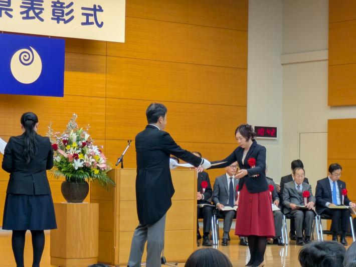 演題の前で正装の男性からスーツ姿の女性に賞状を手渡している