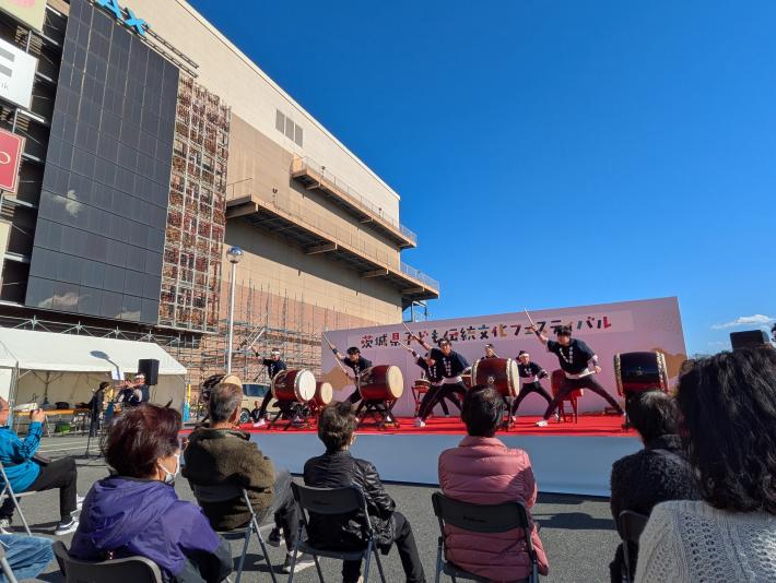 青空の下、特設会場ステージ上で法被姿の人々が、客席に向けて和太鼓を演奏している