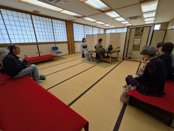 和室に茶席が設けられ、中央に茶をたてる人、両脇に客人が座っている