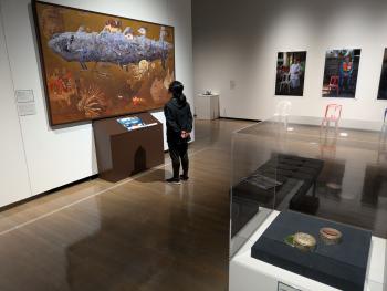 壁や床に美術作品が展示され、壁に掛けられた魚の絵のような作品を鑑賞する一人の人物が映っている