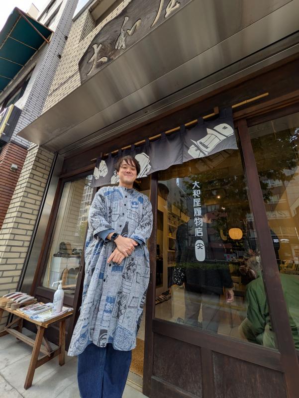 伝統的民具などの絵をあしらった服を着て、下駄屋の店先にたたずむ男性