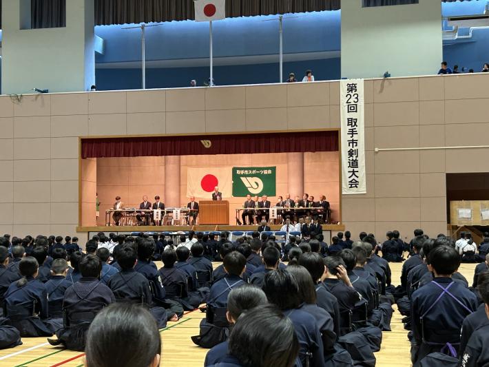 剣道着を着て座る多くの学生の前で、壇上からあいさつをする男性