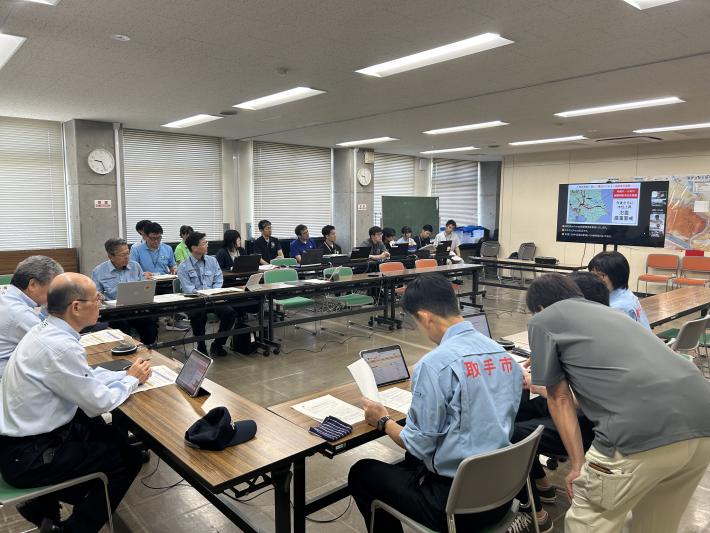 ロの字に並んだテーブルの前に座る防災服を着た人たち