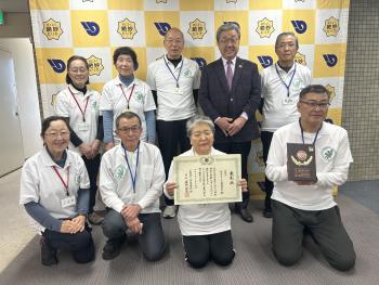 ポロシャツを着た大人8名とスーツを着た大人1名がパネルの前に立っている。