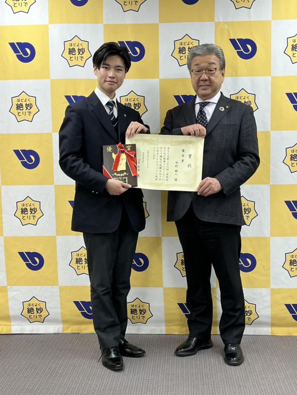 賞状をもつ高校生とスーツを着た男性がパネルの前で立っている