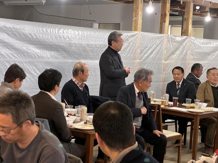 着座の男性に囲まれた男性が立って話をしている