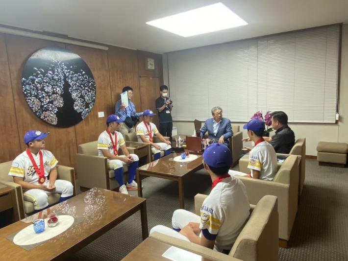 応接いすに腰掛けるスーツの男性と野球ユニフォームを着た中学生たち