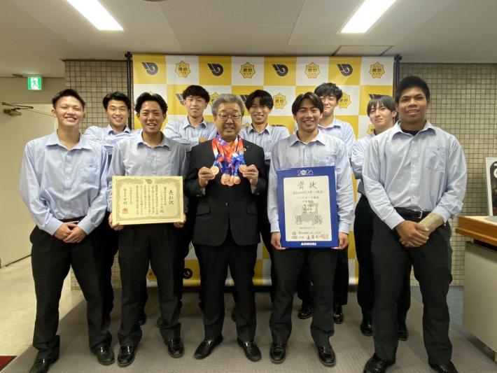 パネルの前に立つ大柄の男子学生たちとスーツを着た男性