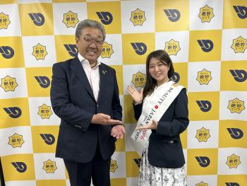 パネルの前に立つ、スーツを着た男性とサッシュをかけた女性