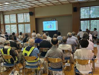体育館の中に座り話を聞く人と前で話をする男性