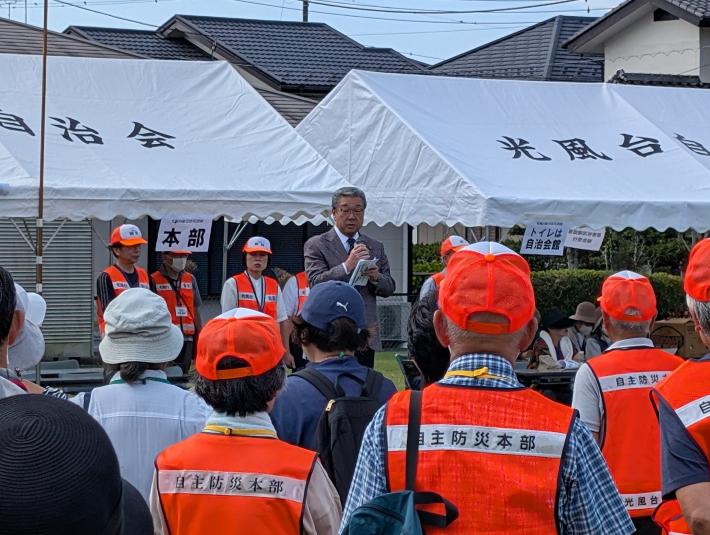 オレンジ色のジャンパーを着た人たちの前からあいさつをする男性