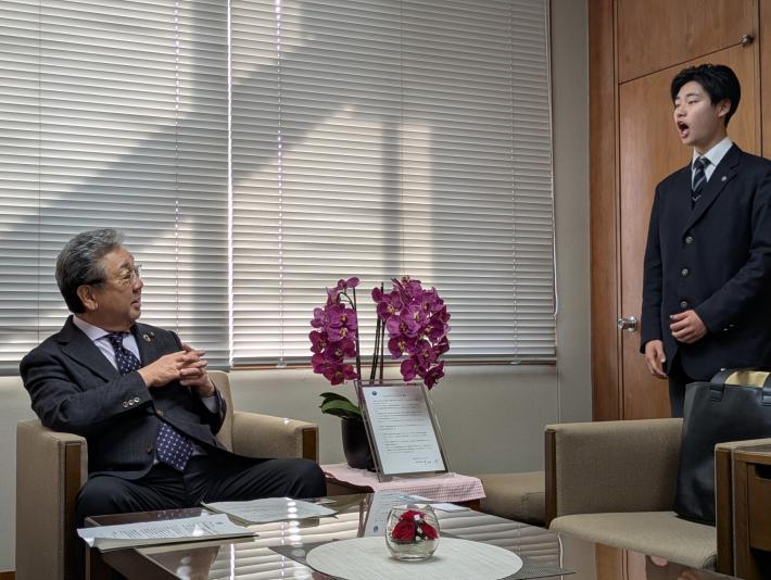 アームチェアに腰かける男性に向けて歌う高校生