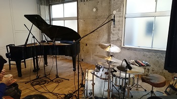 コンクリート打ちっぱなしの壁、板の間にピアノとドラムがおかれている写真