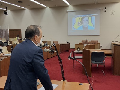 男性が大きくスクリーンに映し出されている。スクリーンの男性に対して質疑席で起立してマイクに向かって質疑する男性議員の写真