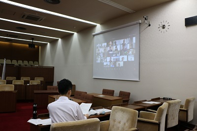 オンライン出席した議員がタイル状に並んでいる画面がスクリーンに映し出されている。男性職員が着座でカメラに向かって説明している写真