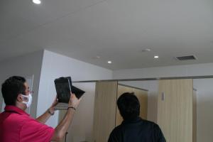黒色の服を着た職員が天井を見ながら説明、ピンク色のポロシャツを着た職員がタブレットを持ちトイレ個室の天井(白色)を写している