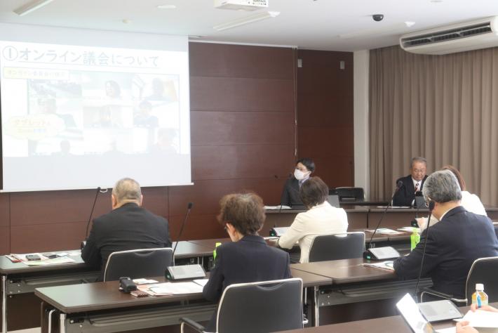 会議室で男性と女性の議員がスクリーンを見ながら話を聞いている