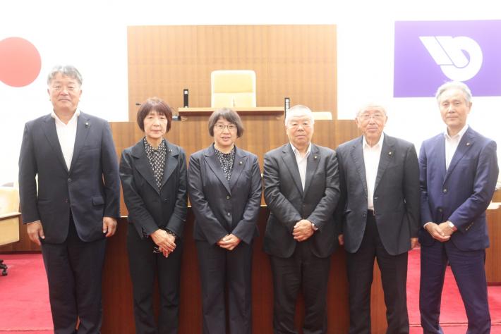 議場で男性と女性の議員数名が立って横に並んでいる