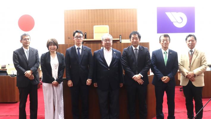 議場で男女数名の議員が立って横に並んでいる