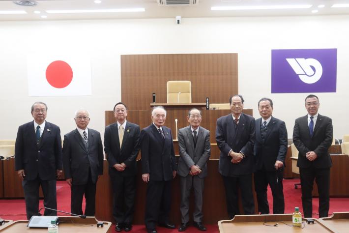 男性の議員数名が議場で並んで立っている