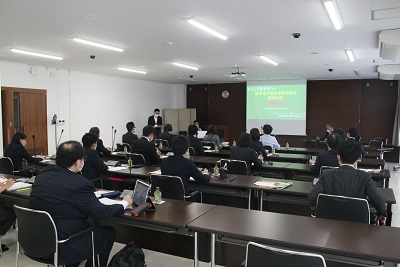 大会議室に机が5列並べられている。緑色の資料が投影されるスクリーン右脇に金澤議長、左脇に立つ司会の職員と座る職員が映る。
