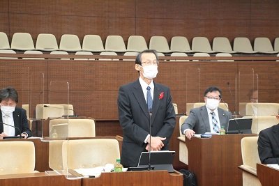 四角いフレームの眼鏡をかけ、青いネクタイを着けた男性議員が議席にて起立し挨拶している。周りには男性議員が3人映る。