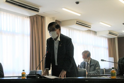 大会議室にて眼鏡をかけた黒色のスーツの男性委員長が起立し挨拶をしている写真。後ろには灰色のスーツを着た男性が映る