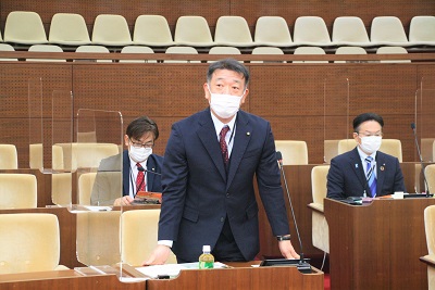 議席最前列中央で起立しあいさつをする赤地に白の水玉模様のネクタイをした男性委員長。後ろには男性市議2人が映る
