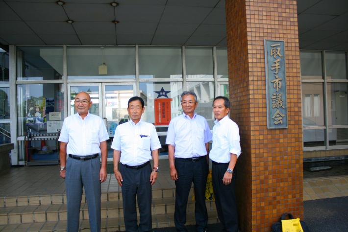 取手市議会と書かれた茶色の柱の横に、白いワイシャツを着た男性4名が横一列に並んで、笑顔でこちらを向いている