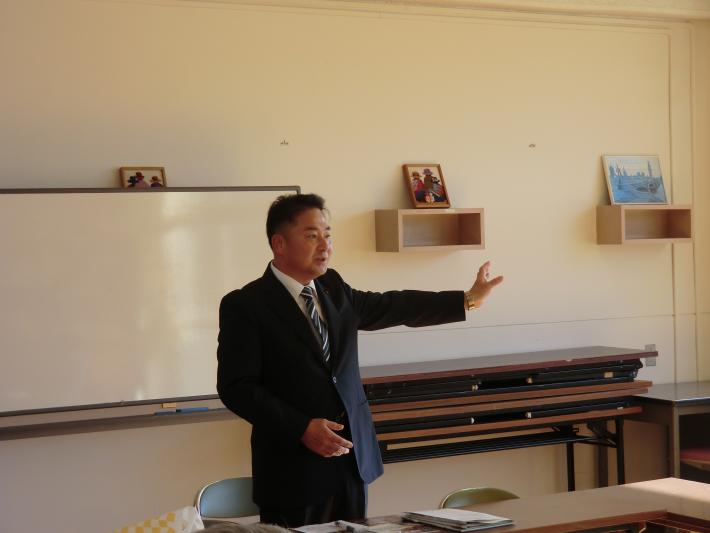 教室の壁にホワイトボードが設置されている。その前に黒いスーツを着た短髪の男性が立っており、左手を上げながら話している