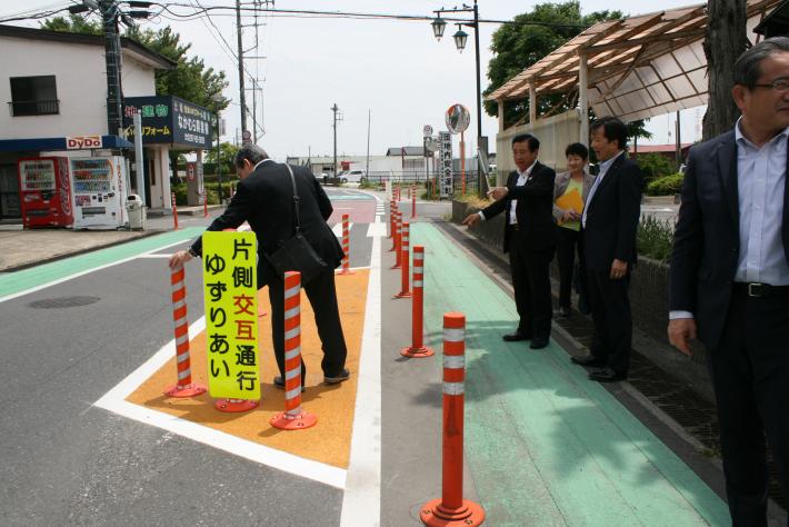 緑色の歩道にスーツを着た男性3人と女性1人が立っている。道に黄色の看板が置かれその脇にスーツを着た男性が立っている