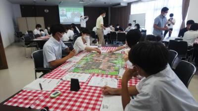 テーブルクロスがひかれた机に生徒4人座っているところに福岡市議1人も加わりボードゲームを用いて対話をしている写真