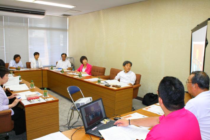 会議室内に、縦長の机4つが長方形に置かれ、ピンクの服を着た男女1名ずつと、白い服を着た男性5名と女性1名が向かい合って座っている
