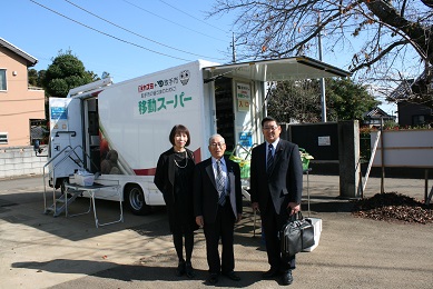 移動スーパーと書かれたトラックを背景に、黒いスーツを着た女性1人と男性2人が笑顔で立っている