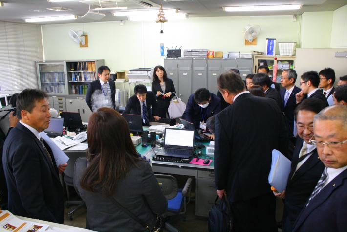 紺色の上着を着た短髪の男性が机上のパソコンを操作している。その男性をスーツを着た男性15人と女性2人が取り囲んでいる