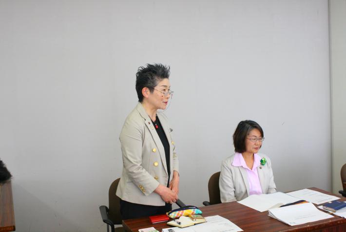 縦長の机が置かれ、灰色のスーツを着た短髪の女性が立っており、隣に白いスーツを着た短髪の女性が着座している